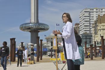 A scientist in a white lab coat speaking next to the i360 on Brighton seafront