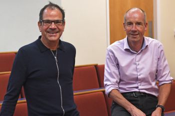 Two men pictured on perching on red seats in a lecture theatre smiling at the camera