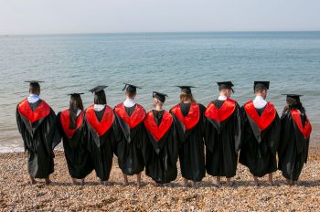 The backs of 9 University of Sussex graduates in gowns on a sunny day on Brighton beach facing the sea.
