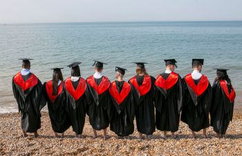 The backs of 9 University of Sussex graduates in gowns on a sunny day on Brighton beach facing the sea.