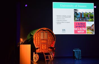 A woman speaks on stage with a screen and props around her