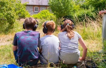 Young people are sitting on the grass wearing headphones and listening to sounds from 'a world never-before encountered' inside a tree with soundscape ecologist, Dr Alice Eldridge.