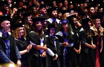 Rows of graduates standing in auditorium, laughing and smiling, wearing graduation gowns and mortar boards