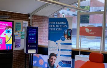 An image showing the new STI screen in a reception area next to a digital screen and a red chair