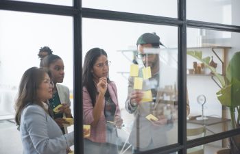 3 women and one man viewed through an office glass pane putting sticky notes on the glass