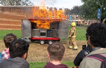 Fire billows from a demonstration kitchen with a fire fighter and students watching