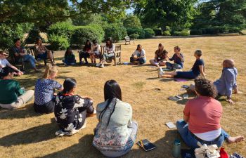 Postgraduate researchers sit in a circle outside on the ground listening to a speaker