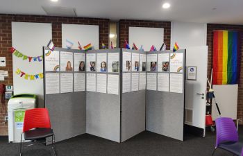 A picture of the LGBTQ+ display featuring the LGBTQ+ notable people in STEM, with a pride flag hanging to the left and bunting that reads 