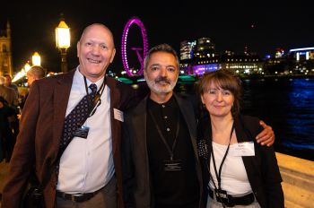 3 people standing on a balcony over the river Thames with the london eye illuminated behind them