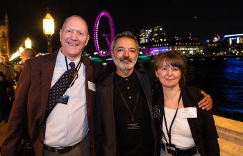 3 people standing on a balcony over the river Thames with the london eye illuminated behind them