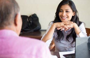 A woman wearing office attire sits behind a desk, smiling while listening to the man sat across from her