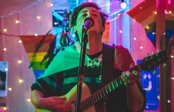 A photograph of a person holding a guitar and singing into a microphone. Behind them is bunting in the Pride flag colours and other decorations and lights.