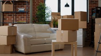 Brown boxes are piled around a partially empty room, with a sofa wrapped in plastic