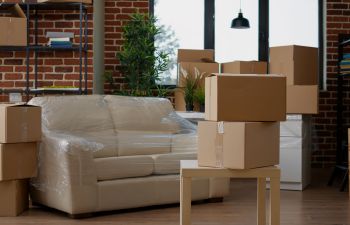 Brown boxes are piled around a partially empty room, with a sofa wrapped in plastic