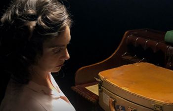 Woman in dramatic lighting, hair up, old briefcase sitting on a desk in front of her.