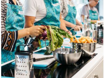Four people stand at a kitchen counter wearing bright blue aprons, they are all chopping vegetables