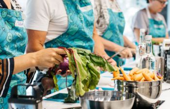 Four people stand at a kitchen counter wearing bright blue aprons, they are all chopping vegetables