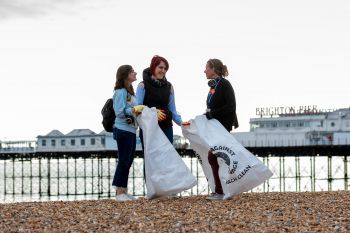 Two students taking part in a beach clean with Kelly Coate, PVC for Education and Students. They are holding white bags with the Surfers Against Sewage Logo.