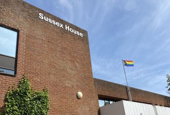 The Pride flag raised above the University of Sussex Building. A blue clear sky and a rainbow coloured flag.