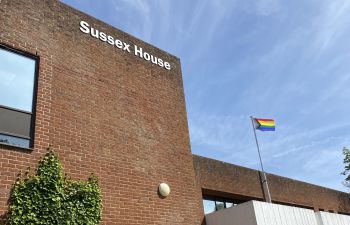 The Pride flag raised above the University of Sussex Building. A blue clear sky and a rainbow coloured flag.