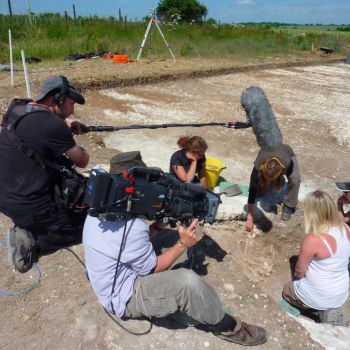 A group of people on a dig site; one is holding a camera, while the other holds a microphone boom. They are filming the excavation.