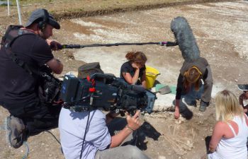 A group of people on a dig site; one is holding a camera, while the other holds a microphone boom. They are filming the excavation.