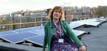 Sam Waugh smiles and sits in front of an array of solar panels, on the roof of Arts C.