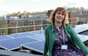 Sam Waugh smiles and sits in front of an array of solar panels, on the roof of Arts C.
