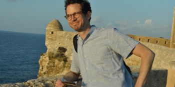 Iain McDaniel standing against a wall, smiling. In the background is the sea and a blue sky.