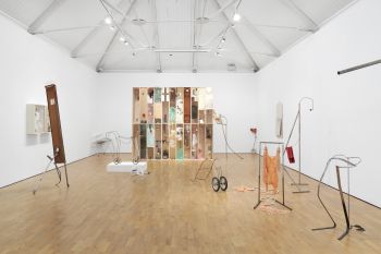A room filled with different objects with contrasting materials: metal sculptures, hanging fabrics and wire frames with wheels attached to them.