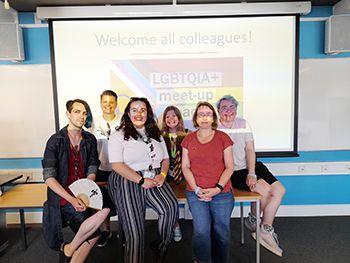 A photo of the members of staff who make up the LGBTQ+ and Trans and Nonbinary Staff Network committees, They are standing in front of a projected image of the LGBTQ+ staff network logo.