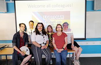 A photo of the members of staff who make up the LGBTQ+ and Trans and Nonbinary Staff Network committees, They are standing in front of a projected image of the LGBTQ+ staff network logo.