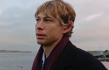 A headshot of Mahon O'Brien on the seafront. He has blonde hair, light skin, and is wearing a scarf.