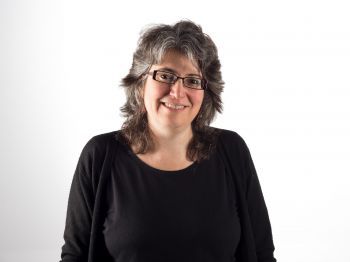 Pam Thurschwell pictured against a white studio background. She is wearing a black top and black glasses, with grey hair.
