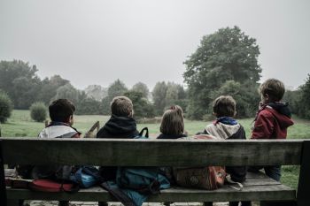 5 children sitting on a bench in a park, facing away from the camera.