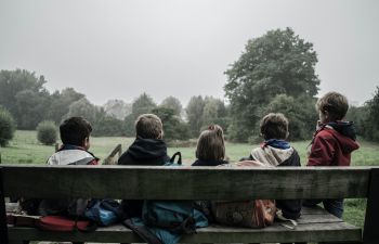 5 children sitting on a bench in a park, facing away from the camera.