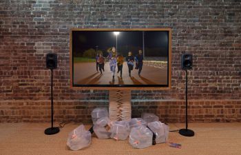 A large TV displaying 6 young adults on a running track, running towards the screen. It’s dark but the track is lit by floodlight.