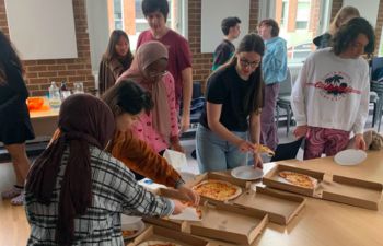 A group of people standing around a table with pizzas, talking with one another.