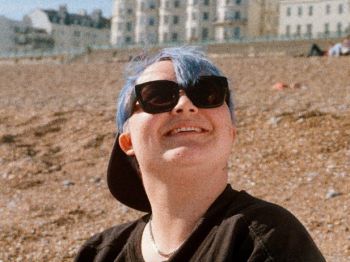 A photo of James who is a non-binary person with a black cap, tshirt and sunglasses on. James is sitting on the beach in Brighton and is smiling.
