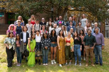 A group of Masters students posing for a group photo. They are standing outside one of the university buildings, underneath a tree.