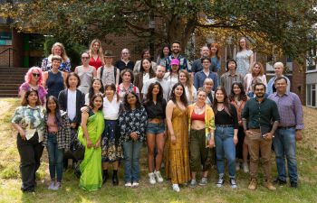 A group of Masters students posing for a group photo. They are standing outside one of the university buildings, underneath a tree.