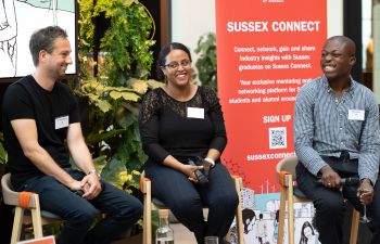 Speakers Omid Moallemi, Emnet Tiruneh and Benjy Kusi smiling as they sit in stools in front of the audience. Behind them is a Sussex Connect banner.