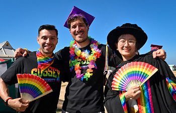 3 people wearing rainbow and black university of sussex t-shirts. Holding rainbow fans in their hands and wearing graduation gowns.