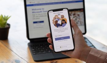 A hand holds a phone showing the Teams app login page. Behind their hand is an out of focus image of a laptop screen.