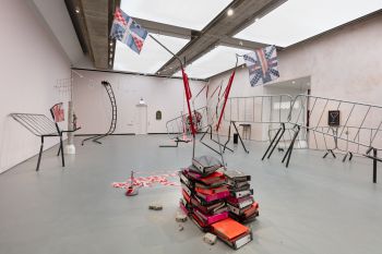 A room filled with contrasting objects; warped metal barriers and structures stand elevated, a pile of orange, pink and black books, a warped ladder and structures with White, Red and Blue fabric resembling the Union Jack flag.