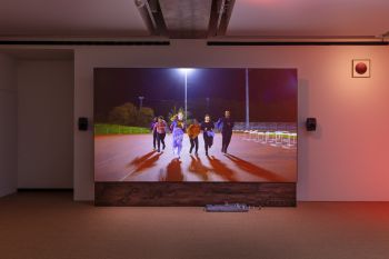 A large TV displaying 6 young adults on a running track, running towards the screen. It’s dark but the track is lit by floodlight. They are wearing black and bright tie died clothing.