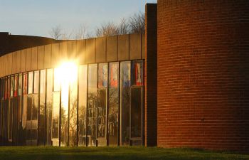 A photo of outside of the ACCA building on a clear blue sky sunny day. To the right third of the shot the curved wall of the building has orange bricks. The left two thirds of the shot feature the glass wall with the sun reflecting off it.