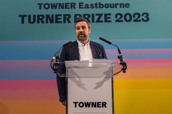 A photo of Joe Hill, the director of Towner Eastbourne. He stands behind a podium wearing a white shirt and blue suit. Behind him is a large rainbow banner with “Turner Prize 2023” printed across it.