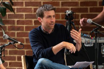 An action photo of Ben Burbridge during an interview. He is sitting on a black chair with a brick wall behind him. He is surrounded by audio and visual equipment.