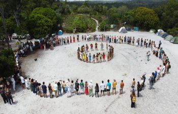 a circle of people pictured standing in the sand and listening to indigenous leaders in the middle of the circle